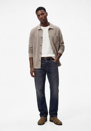 Mann trägt beiges, strukturiertes Hemd mit Knöpfen, weißes T-Shirt, dunkelblaue Jeans, braunen Gürtel und braune Sandalen mit geschlossener Spitze, steht mit einer Hand in der Hosentasche.