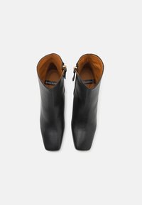 Bottes chevilles en cuir noir avec une texture lisse, bout carré et accents de fermeture éclair dorés. L'intérieur présente une doublure brune contrastante.
