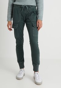 Persona que lleva pantalones cargo joggers verde oscuro con bolsillos frontales y en los muslos, combinados con zapatillas blancas y un suéter de canalé gris claro.