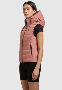 Padded bezrukávový vestník v jemnej ružovej farbe s vysokým golierom a veľkou kapucňou. Obsahuje bočné zipsové vrecká a elastické bočné panely pre pohodlie.