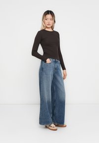 Langermet brun topp kombinert med vide blå denimjeans. Jeansene har en falmet vask med slitt nederkant, og skoene er beige med et mønster.