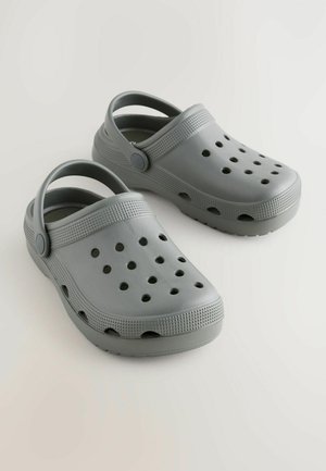 Pelēki gumijas klapki ar perforētu dizainu, aizmugures siksnu, teksturētām joslām un apaļu formu; piemēroti ikdienas valkāšanai un vieglai tīrīšanai.