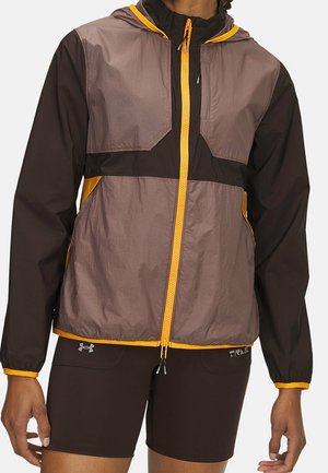 Person trägt eine leichte braun-gelbe Kapuzenjacke und schwarze Under Armour Shorts, steht mit entspannten Armen an den Seiten.