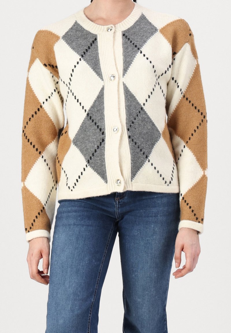 Femme portant un cardigan boutonné avec un motif losanges beige, gris et blanc, associé à un jean bleu, debout avec les bras détendus le long du corps.