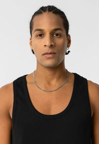 Zwarte geribbelde tanktop gedragen door een persoon, met een zilveren kettinghalsband. De achtergrond is eenvoudig wit en textuur. 