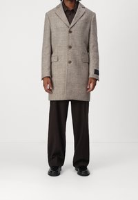Abrigo de cuadros gris con solapa muesca, tres botones, bolsillos laterales y una etiqueta de tela negra, combinado con pantalones marrones y zapatos negros.