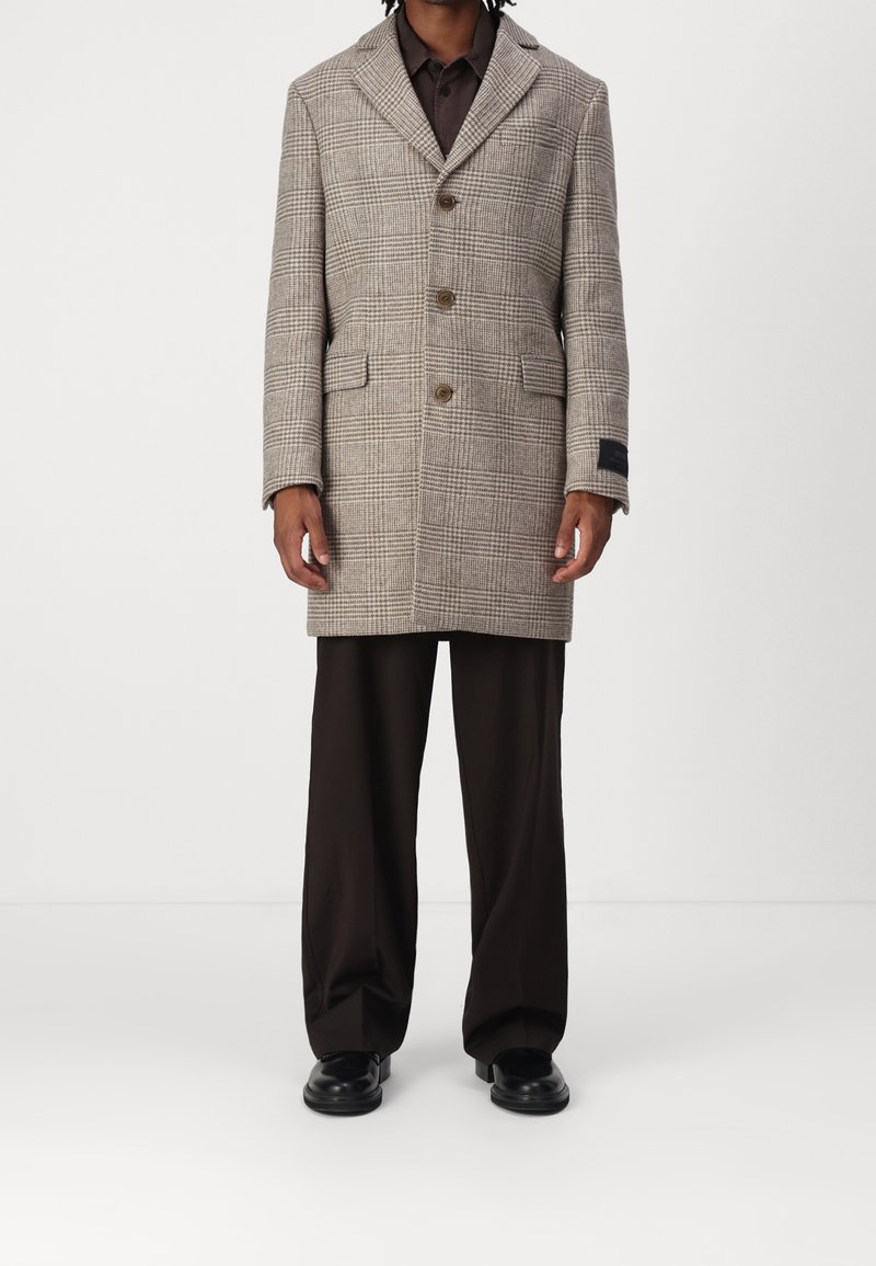 Abrigo de cuadros gris con solapa muesca, tres botones, bolsillos laterales y una etiqueta de tela negra, combinado con pantalones marrones y zapatos negros.