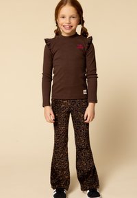 Bruine lange mouw top met ruches, gecombineerd met flared broek in luipaardprint. Model draagt zwarte sneakers. Neutrale achtergrond.