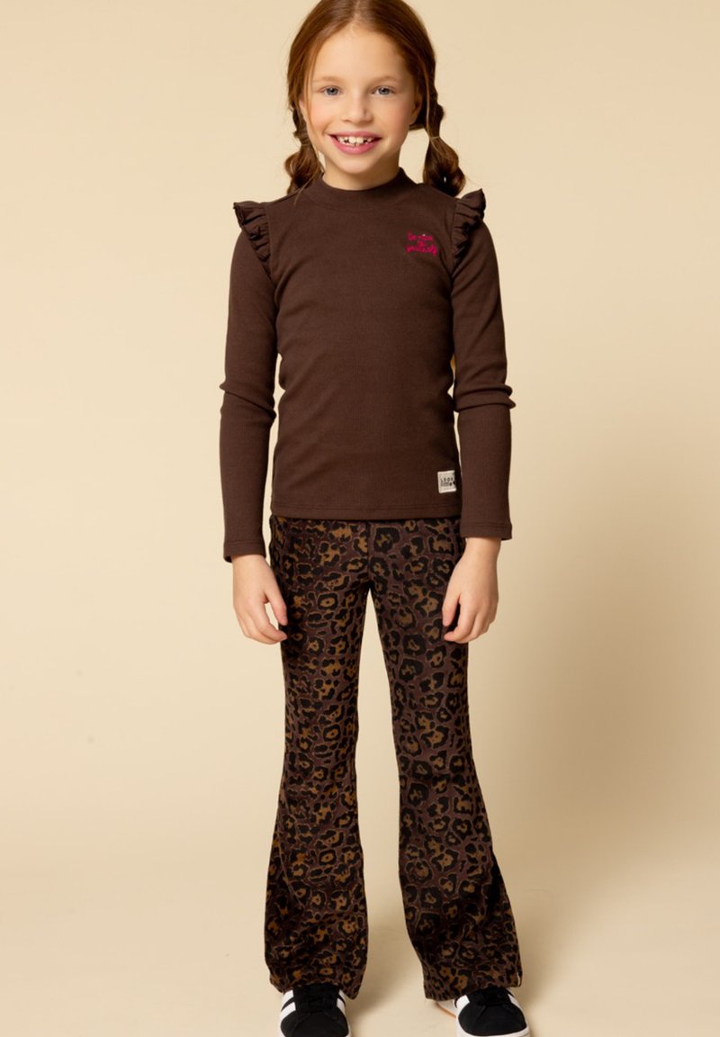 Bruine lange mouw top met ruches, gecombineerd met flared broek in luipaardprint. Model draagt zwarte sneakers. Neutrale achtergrond.