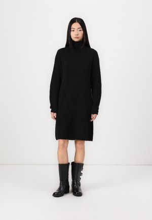 Black knit sweater dress with a high collar, long sleeves, and a straight cut. Worn with knee-high black boots featuring side buckles.