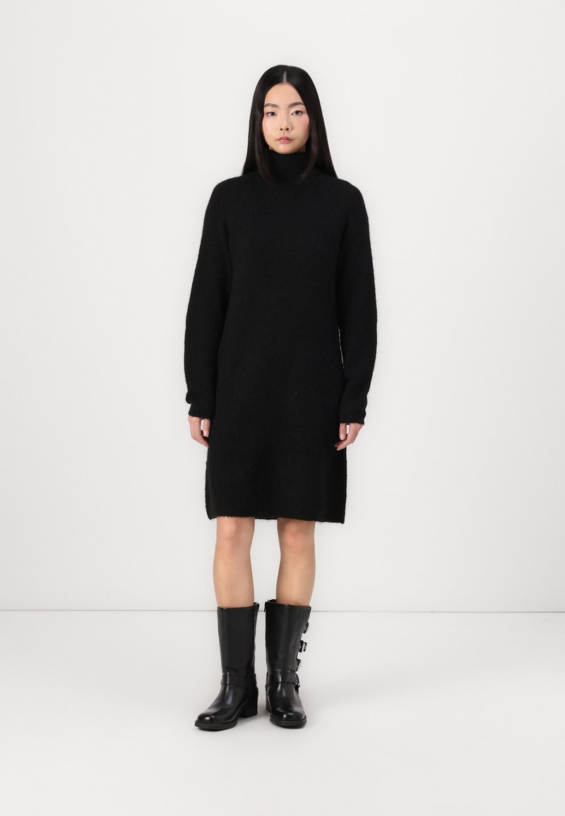 Black knit sweater dress with a high collar, long sleeves, and a straight cut. Worn with knee-high black boots featuring side buckles.