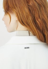 Blazer bianco con colletto strutturato, caratterizzato da una spilla con il logo "BOSS" in argento sul retro, completato da un tessuto beige testurizzato sotto.