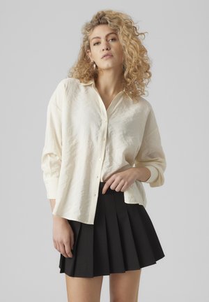 Chemise blanche en lin oversize à boutons et manches longues, associée à une mini-jupe plissée noire. Texture lisse, coupe décontractée et détails minimaux.