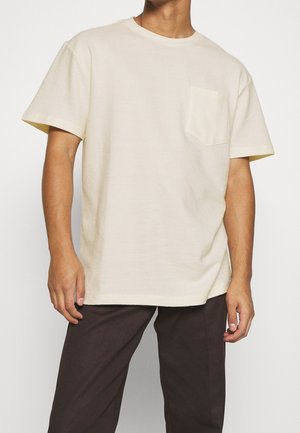Man wearing a plain off-white short-sleeve T-shirt with a chest pocket and dark brown pants, standing against a white background.