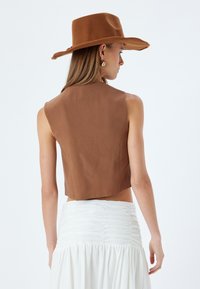 Gilet corto marrone in tessuto leggero, con scollatura pulita e design sul retro aperto. Abbinato a una gonna bianca a volant. Cappello marrone a tesa larga.