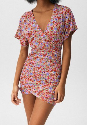 Femme portant une robe courte et ajustée à fleurs rouges, violettes et roses, avec un décolleté en V, sur un fond uni.