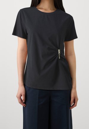 Femme portant un top noir à manches courtes avec des fronces sur les côtés et un petit détail chaîne dorée, associé à un pantalon large foncé.