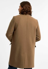 Brązowy płaszcz z wełny z domieszką, o dopasowanym kroju, z wyraźnymi ramionami i bocznymi kieszeniami. Gładka faktura i czyste linie, widok z tyłu.