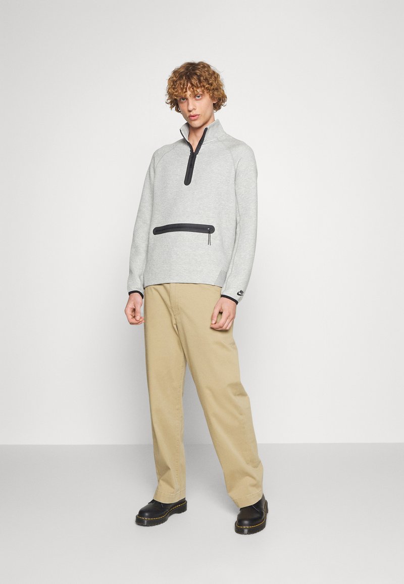 Pull-over en polaire gris clair avec un zip demi-long, poche poitrine zippée noire, et accent noir au col ; associé avec un pantalon beige et des chaussures noires.