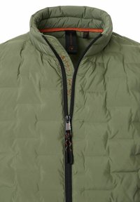 Leichte olivgrüne Pufferjacke mit Stehkragen, schwarzem Reißverschluss und strukturierten Oberflächen. Verfügt über rote gedruckte Koordinaten in der Nähe des Reißverschlusses.