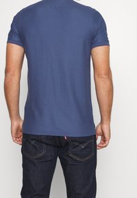 Modrá bavlněná tričko s krátkými rukávy, kulatým výstřihem a rovním lemem. Nosič má na sobě tmavé džíny s kontrastním šitím.