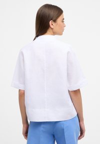Blusa bianca a maniche corte in lino, con una silhouette pulita, dettagli sulla cucitura posteriore e polsini leggermente ripiegati.