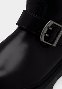 Κοντινό πλάνο λαμπερού μαύρου δερμάτινου μποτάκι με ραμμένο λουράκι και ασημένια μεταλλική αγκράφα χαραγμένη με το όνομα της μάρκας.