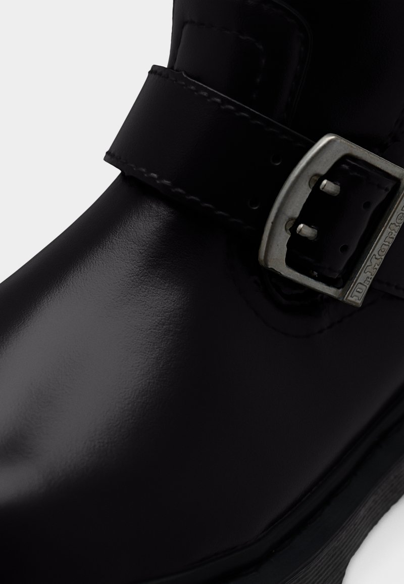 Primo piano di uno stivale in pelle nera lucida con cinturino cucito e fibbia in metallo argentato incisa con il nome del marchio.