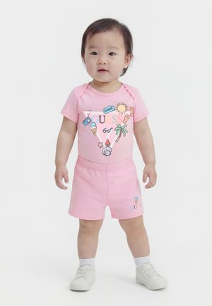 Tout-petit debout, portant une tenue d'été rose avec des motifs tropicaux et de glace, des chaussettes blanches et des baskets blanches sur un fond uni.