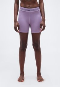 Άτομο στέκεται ξυπόλητο φορώντας λιλά αθλητικές βερμούδες με ψηλή μέση και λογότυπο New Balance σε απλό λευκό φόντο.