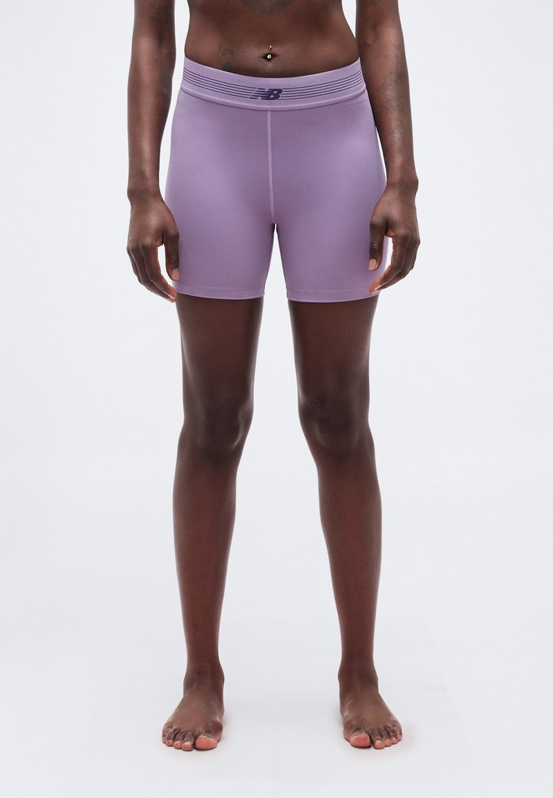 Άτομο στέκεται ξυπόλητο φορώντας λιλά αθλητικές βερμούδες με ψηλή μέση και λογότυπο New Balance σε απλό λευκό φόντο.