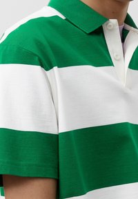 Henkilö, jolla on päällään vihreä-valkoisiraidallinen t-paita, jossa on lyhyet hihat ja kaksi nappia.