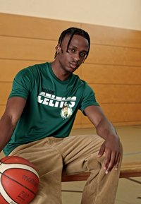 Zöld, rövid ujjú póló, rajta a "Boston Celtics" logóval; világosbarna kordbársony nadrág; kosárlabda a padlón; fa tornaterem háttér.