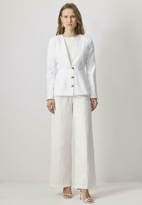 Valkoinen pellavapuvun takki kultaisilla napeilla ja V-kaula-aukolla, yhdistettynä leveälahkeisiin valkoisiin pellavahousuihin. Sulava pinta, minimalistinen muotoilu, elegantti siluetti.