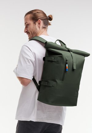 Sac à dos à rabat vert avec des bretelles ajustables, fermeture éclair latérale et étiquette colorée. Fabriqué en matériau durable, il présente un design épuré.