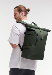Mochila de tipo roll-top verde, fabricada en material impermeable, con correas ajustables, un bolsillo frontal con cremallera y acentos de tiradores de cremallera coloridos.