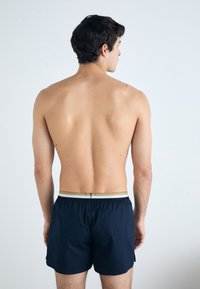 Shorts de bain bleu marine avec une ceinture élastique et une accent en or. Le tissu semble léger avec une texture lisse.