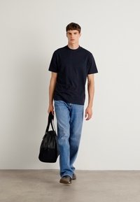 Svart t-skjorte med rund hals i kombinasjon med lys blå jeans og brune sko. Bærer en svart treningst bag, mot en nøytral bakgrunn.