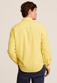Achteraanzicht van een geel linnen overhemd met een kraag, knoopdetails en een gebogen zoom, met een klein logo nabij de hals. Glad van textuur.