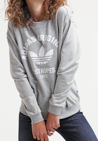 En person som bär en grå Adidas Originals sweatshirt med vitt logotyp och text, i kombination med mörkblå jeans, stående mot en vit bakgrund.
