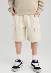 Beige cargo shorts made of cotton blend fabric, featuring two side pockets, drawstring waist, and a relaxed fit. Paired with black sneakers.