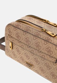 Brązowa torebka na ramię z wzorem, złotymi okuciami, podwójnymi suwakami i przednią kieszenią. Materiał wydaje się teksturowany, ozdobiony detalami z logo na całej powierzchni.