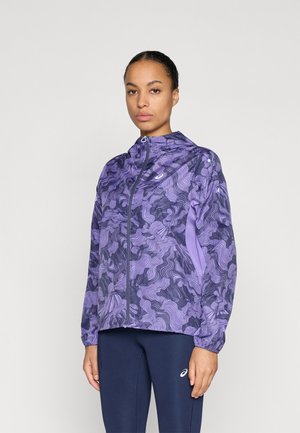 Fialová bunda so zakriveným čiernym vzorom, predný zips a nastaviteľná kapucňa. Ľahký materiál s elastickými manžetami. Kombinovaná s tmavými legínami.