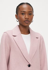 Ροζ ανοιχτόχρωμο παλτό από μείγμα μαλλιού με δομημένο γιακά, κούμπωμα με κουμπιά μπροστά και υφή επιφάνειας. Το μοντέλο φοράει λευκό εσώρουχο.