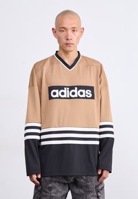 Νεαρός άνδρας με ξυρισμένο κεφάλι, φορώντας αθλητική μπλούζα Adidas σε μπεζ και μαύρο χρώμα με λογότυπο, που στέκεται μπροστά σε λευκό φόντο.