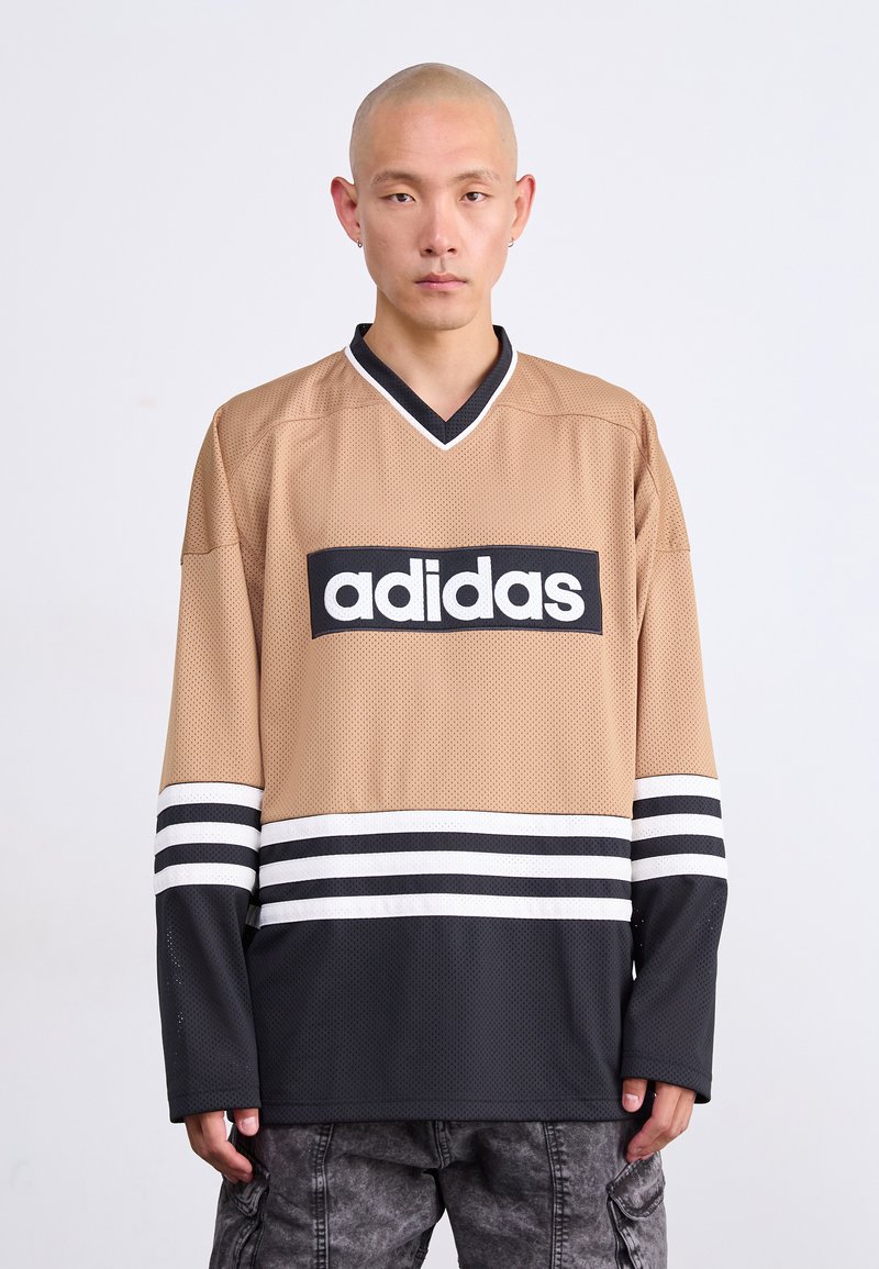 Νεαρός άνδρας με ξυρι�σμένο κεφάλι, φορώντας αθλητική μπλούζα Adidas σε μπεζ και μαύρο χρώμα με λογότυπο, που στέκεται μπροστά σε λευκό φόντο.