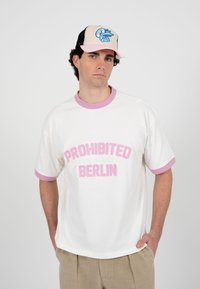 Mann trägt ein weißes T-Shirt mit pinkem Rand und dem Schriftzug "PROHIBITED BERLIN", beige Hose und beige-pinken Hut mit blauem Logo, steht mit den Händen in den Taschen.