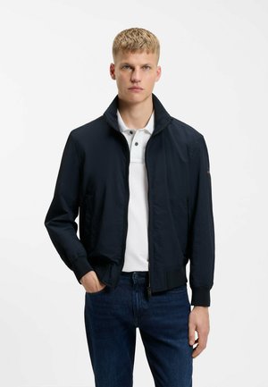 Młody mężczyzna o krótkich blond włosach, ubrany w granatową kurtkę na zamek, na białej koszulce polo i ciemnoniebieskich dżinsach, stoi z jedną ręką w kieszeni.