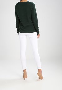Dunkelgrüner strukturierter Pullover, kombiniert mit weißen Skinny-Jeans und nudefarbenen High Heels. Der Pullover hat lange Ärmel und einen runden Ausschnitt.
