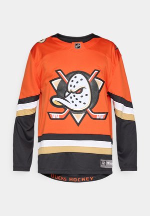 Orangenes Hockey-Trikot mit langen Ärmeln, das in der Mitte ein großes schwarz-weißes Logo aufweist, sowie schwarze und goldene Streifen am unteren Ende.
